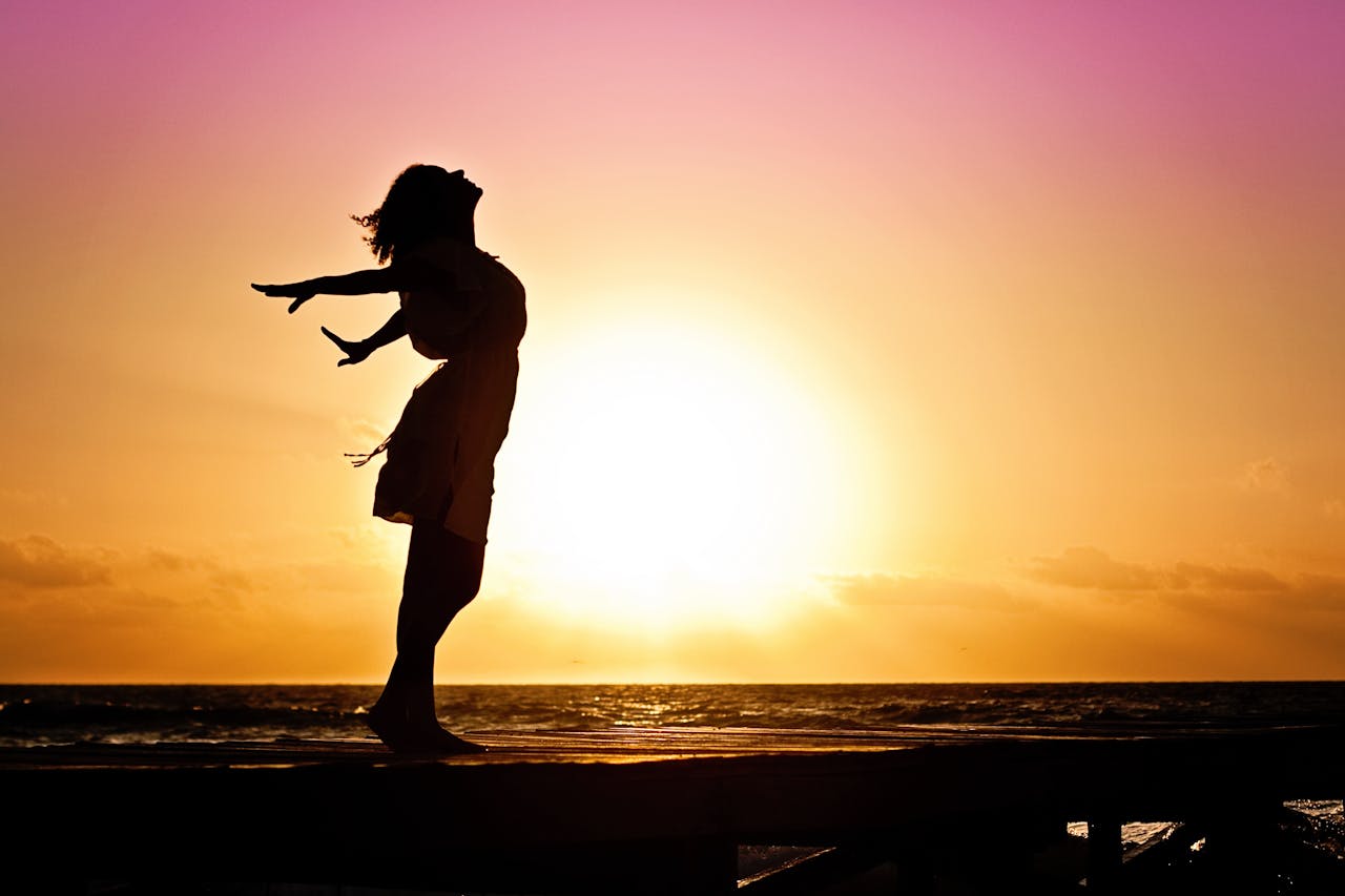 Services Silhouette of a woman at the beach with arms outstretched against a vibrant sunset backdrop.